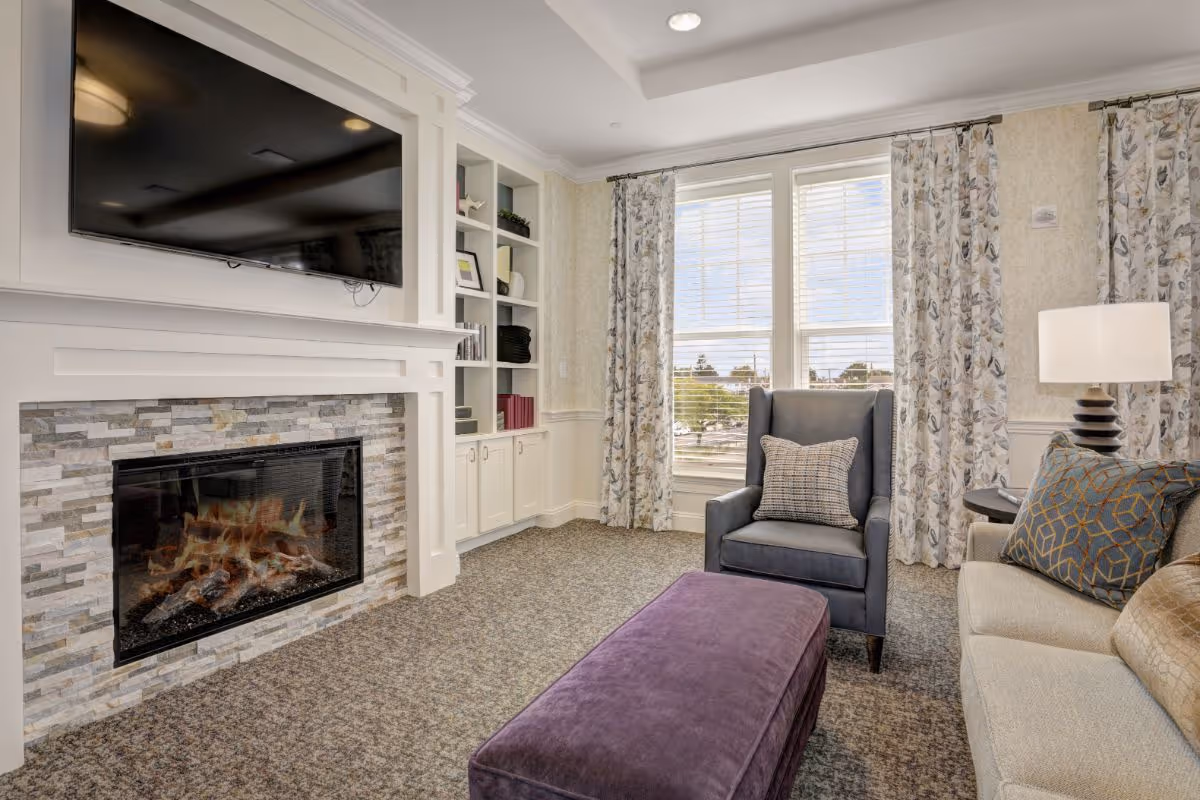 Bright living room with a fireplace and TV, built-in shelving, armchair, sofa, and purple ottoman by a large window.