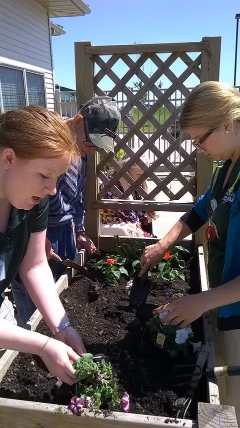 Two women and a man are gardening together outdoors, planting flowers in a raised garden bed filled with soil. A wooden lattice fence is visible behind them, and a person in a wheelchair is seen in the background.
