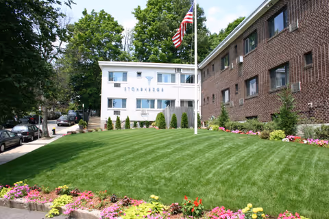 Front lawn and landscaped entrance of Stonehedge Rehabilitation and Skilled Care Center with a white and brick building and an American flag.