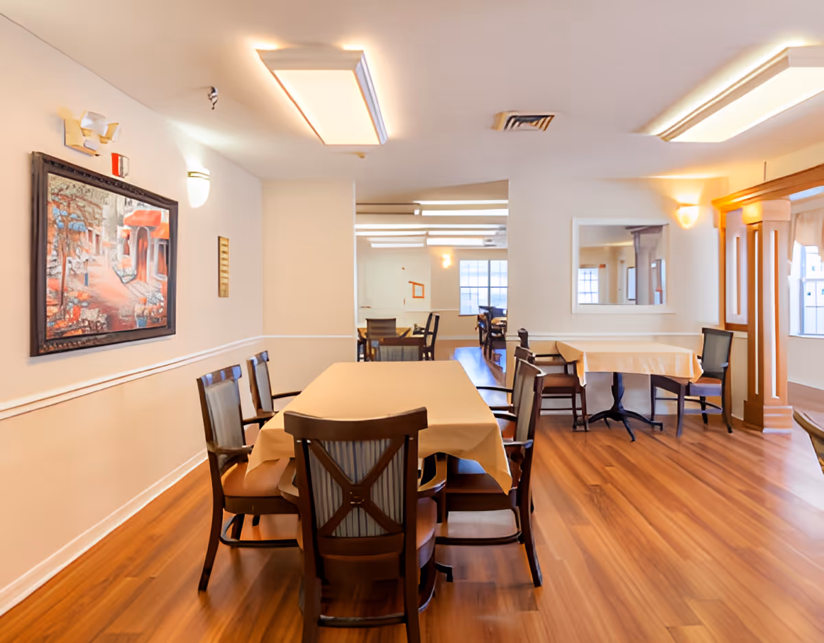 A dining room with wooden floors and multiple tables covered with beige tablecloths. Several wooden chairs with cushioned seats are arranged around the tables. The room is well-lit with ceiling lights and wall sconces. A framed painting and a mirror hang on the walls, and large windows allow natural light to enter.
