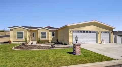 Single-story yellow house with a three-car garage, a well-maintained green lawn, and a small landscaped area with plants near the front entrance under a clear blue sky.