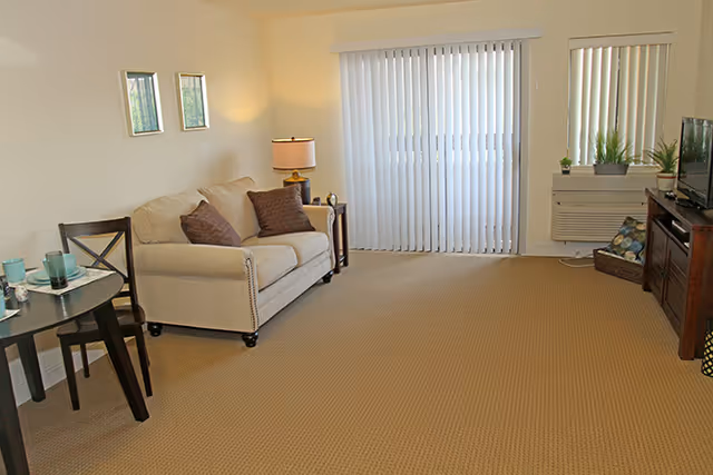 A living room with a beige sofa with two brown pillows, a side table with a lamp, a small round dining table with two chairs, a TV on a wooden stand, and vertical blinds covering a sliding glass door and a window.