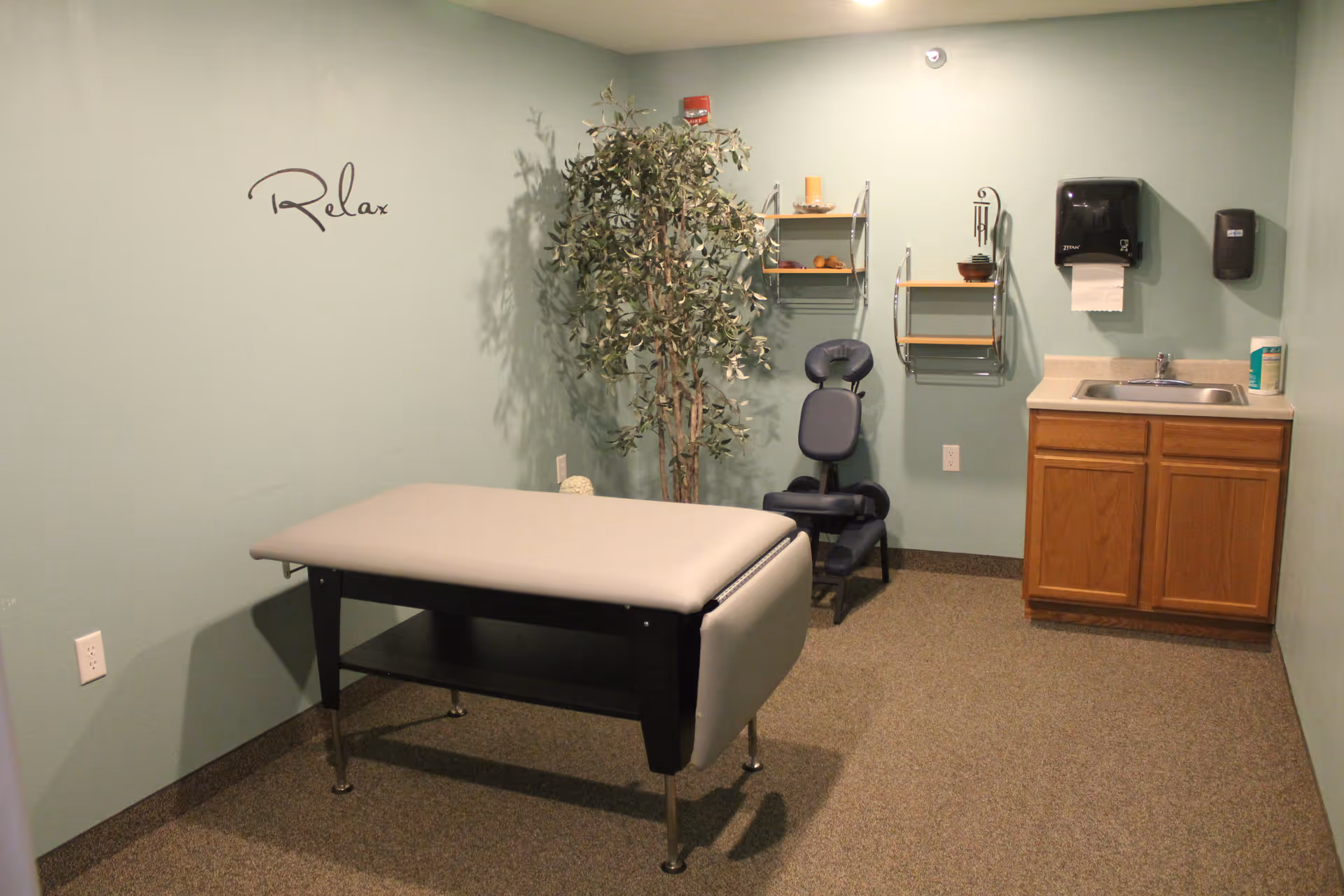 A small therapy or massage room with a padded massage table in the center, a black massage chair against the wall, a tall artificial plant, and a sink with wooden cabinets below. The walls are painted light green and have the word 'Relax' written on one side. There are also two small wall shelves holding decorative items and a paper towel dispenser above the sink.
