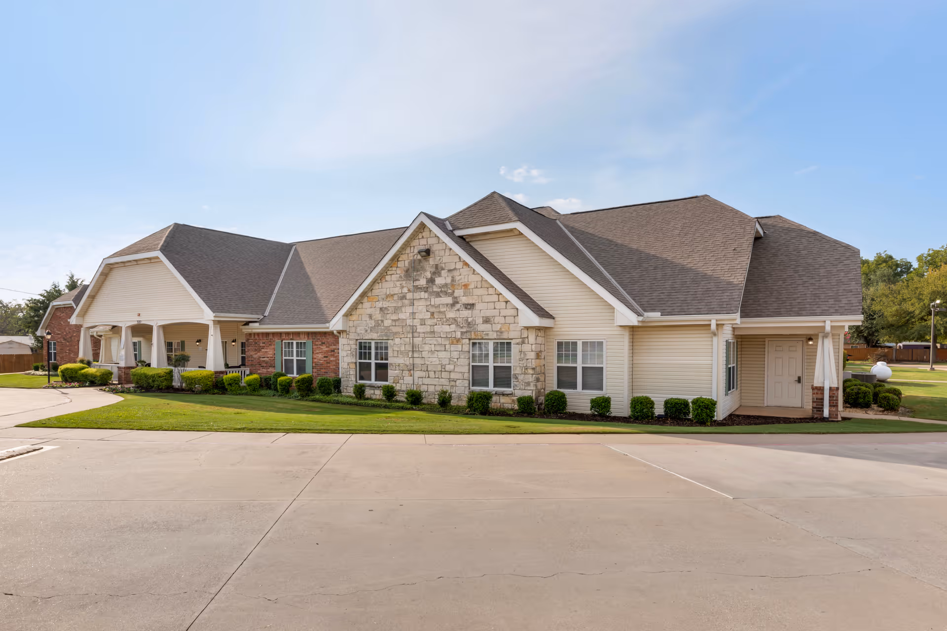Exterior view of a single-story senior living facility building with a combination of stone and siding walls, multiple windows, and a covered entrance. The building is surrounded by neatly trimmed bushes and a well-maintained lawn under a clear blue sky.