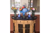 A wooden cabinet with a black countertop placed against a wall with windows covered by white blinds. On top of the cabinet are four glass vases filled with colorful flowers including blue hydrangeas, orange, pink, and yellow blooms. There are also two small blue and white decorative items on the countertop.