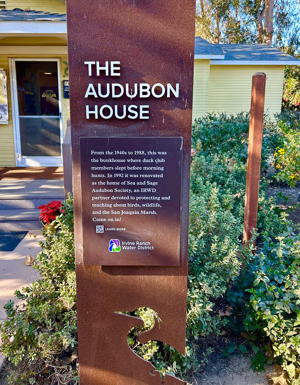 A brown metal sign reading 'THE AUDUBON HOUSE' with a historical plaque stands in front of a yellow building surrounded by plants.