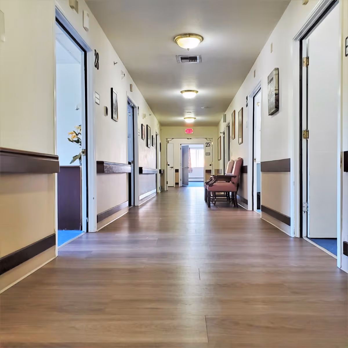A long, well-lit hallway in a senior living facility with wooden flooring and cream-colored walls. Several open doors line both sides of the hallway, with room numbers visible. Two pink cushioned chairs are placed against the right wall near the end of the corridor. Ceiling lights illuminate the space, and framed pictures hang on the walls.