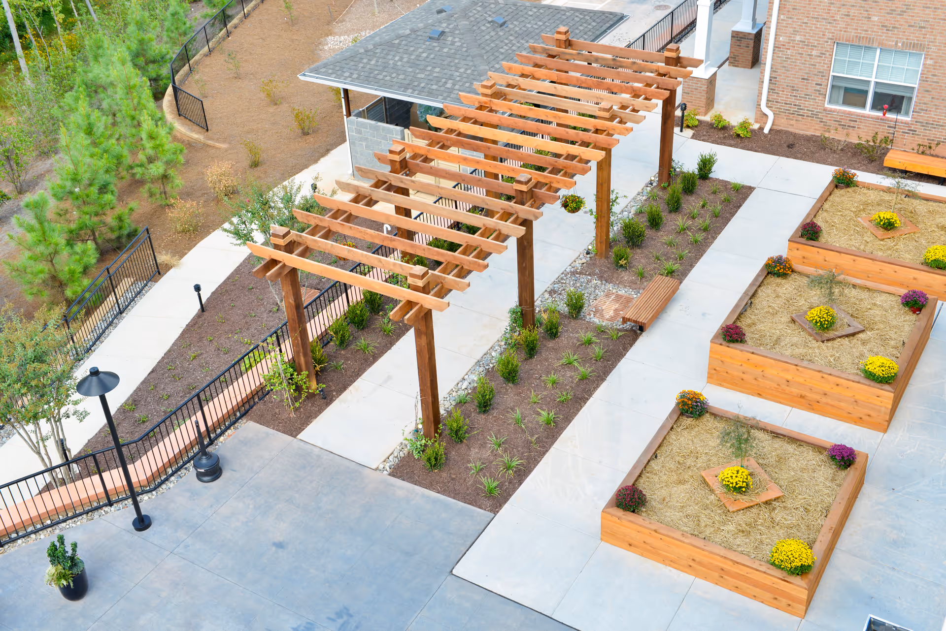 Aerial view of an outdoor garden area at a senior living facility featuring a wooden pergola over a concrete walkway, raised wooden garden beds with flowers and straw, benches, and surrounding landscaping with trees and shrubs.