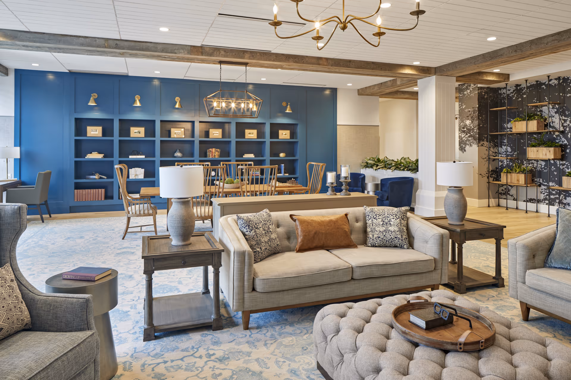 Comfortable communal living room with sofas, armchairs, a tufted ottoman, wooden side tables, and a blue built-in bookshelf and dining area in the background.
