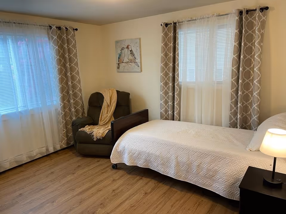 A cozy bedroom with a single bed covered in a white quilt, a small black nightstand with a lit lamp, a comfortable dark armchair draped with a beige throw blanket, two windows with white blinds and patterned beige curtains, and a wall painting of two birds on a branch.