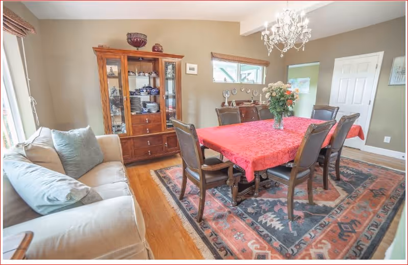 A dining room with a rectangular wooden table covered with a red tablecloth, surrounded by six dark wooden chairs with leather seats. A vase with flowers is placed in the center of the table. To the left, there is a beige sofa with cushions. Behind the table, a wooden cabinet with glass doors displays dishes and decorative items. The room has wooden flooring and a colorful patterned rug under the table. A chandelier hangs from the ceiling, and there is a window with a wooden blind on the wall.