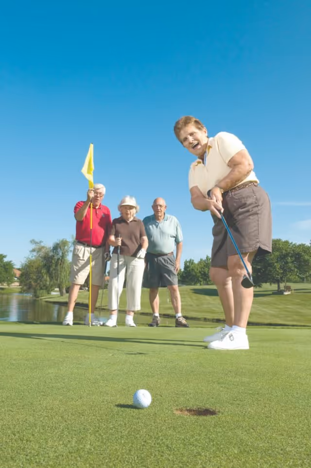 Four elderly people playing golf on a sunny day. One woman is putting the golf ball towards the hole while three others watch, one holding the flagstick. They are on a well-maintained green with trees and a pond in the background under a clear blue sky.