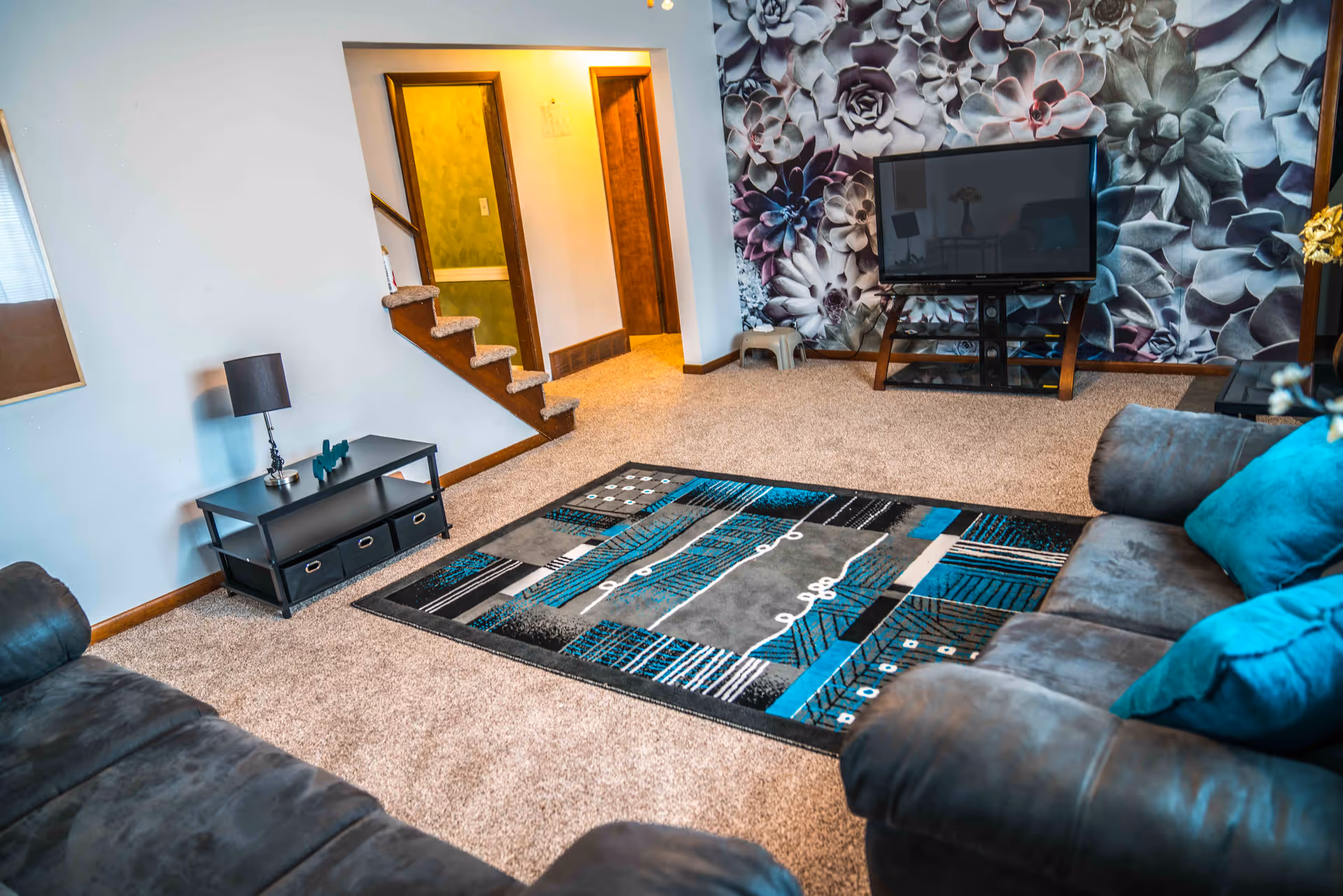 A cozy living room with beige carpet, a patterned area rug in shades of blue, gray, and black, two dark gray sofas with blue cushions, a flat-screen TV on a stand against a wall with large floral wallpaper, a small black side table with a lamp, and a staircase leading upstairs with two doorways visible.