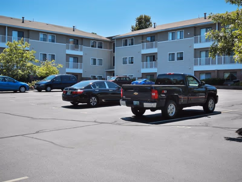 Parking lot in front of a three-story senior living facility building with several parked cars and trees on a sunny day.