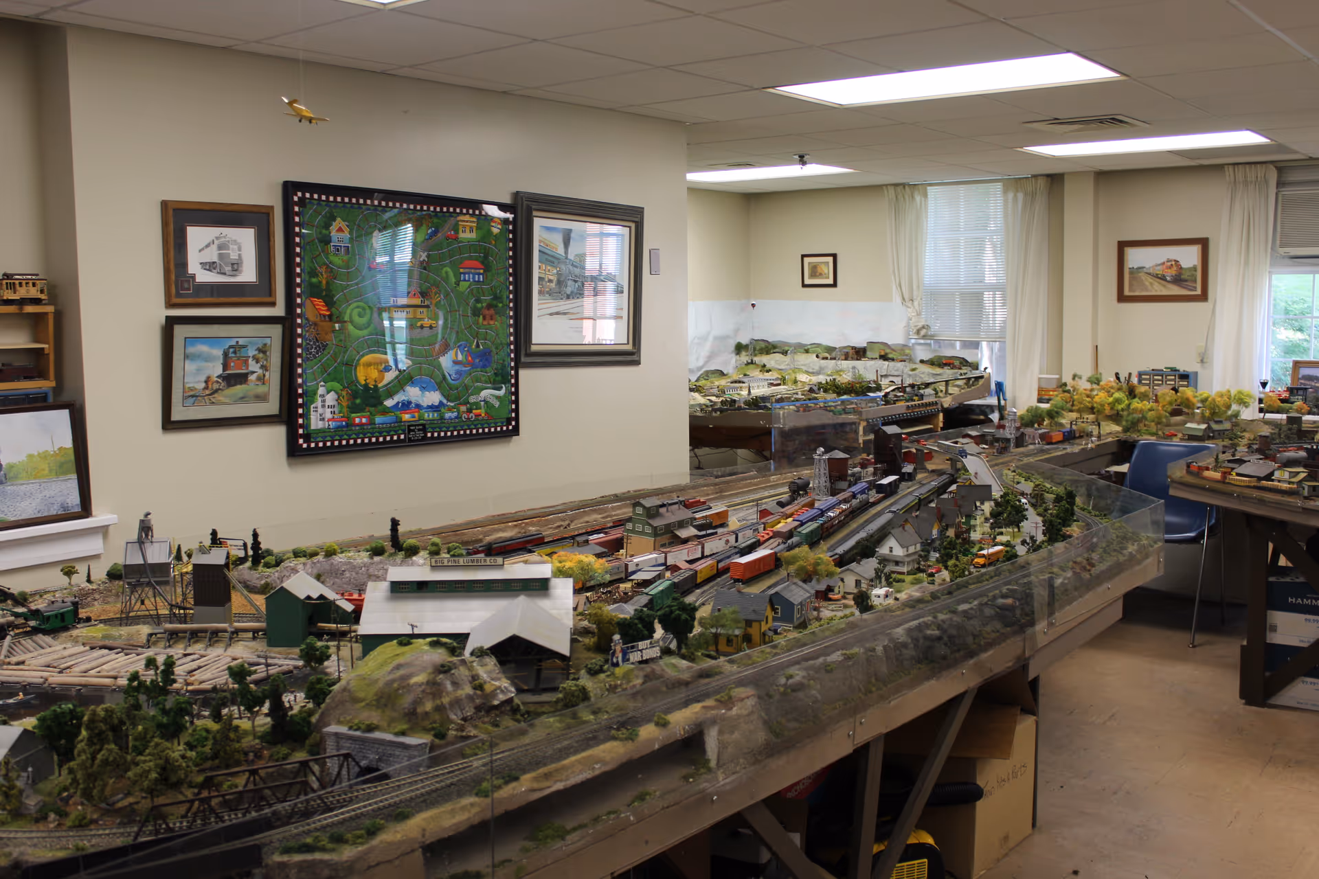 A room with a large model train layout featuring miniature buildings, trees, and trains. The walls have framed pictures and artwork related to trains. The room is well-lit with fluorescent ceiling lights and has windows with white curtains.