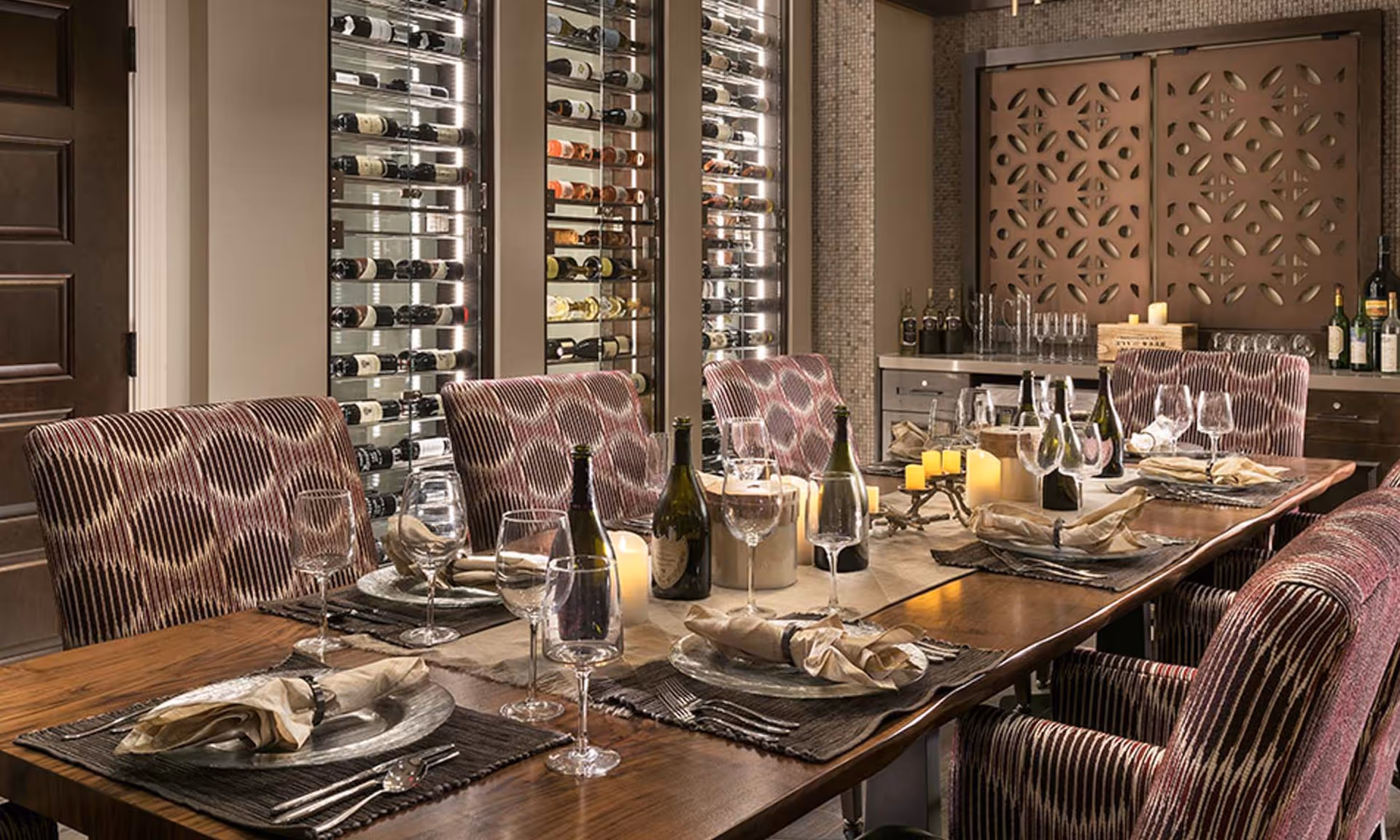 Elegant dining room with a long set table, upholstered chairs, candles, wine bottles and glassware in front of illuminated wine racks.