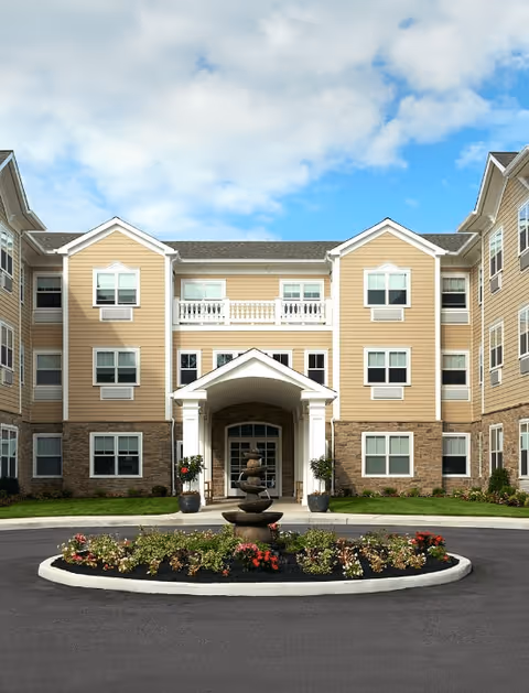 Front exterior view of a three-story senior living facility named Amber Court with beige siding and stone accents. The entrance features a covered porch with white columns, and there is a circular driveway with a landscaped island in the center containing a tiered fountain and flowers. The sky is partly cloudy.