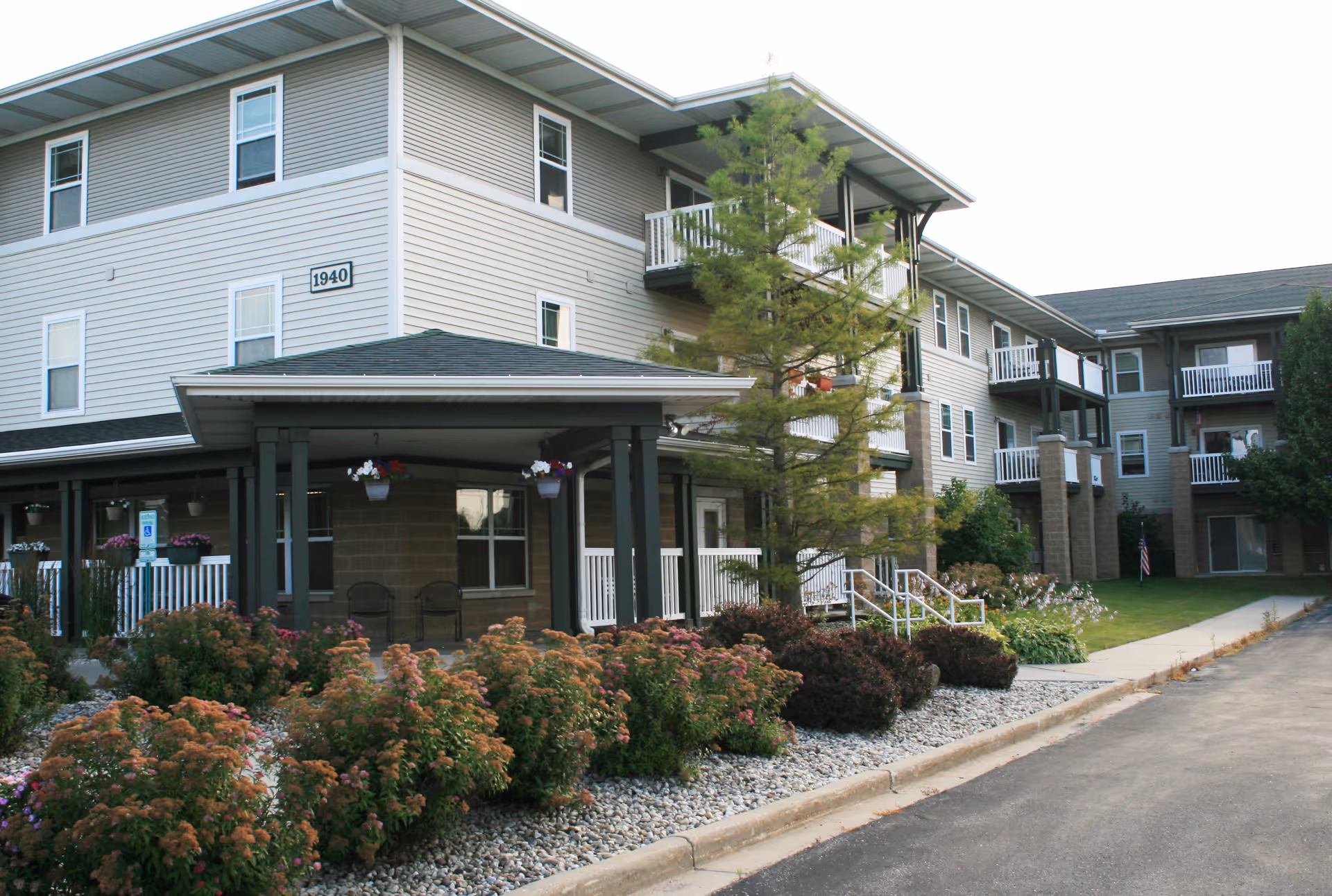 Exterior view of a multi-story senior apartment building with beige siding and white balconies. The building is surrounded by landscaped bushes, flowers, and a small tree. There is a covered porch area with chairs and hanging flower pots near the entrance. A paved driveway runs alongside the building.