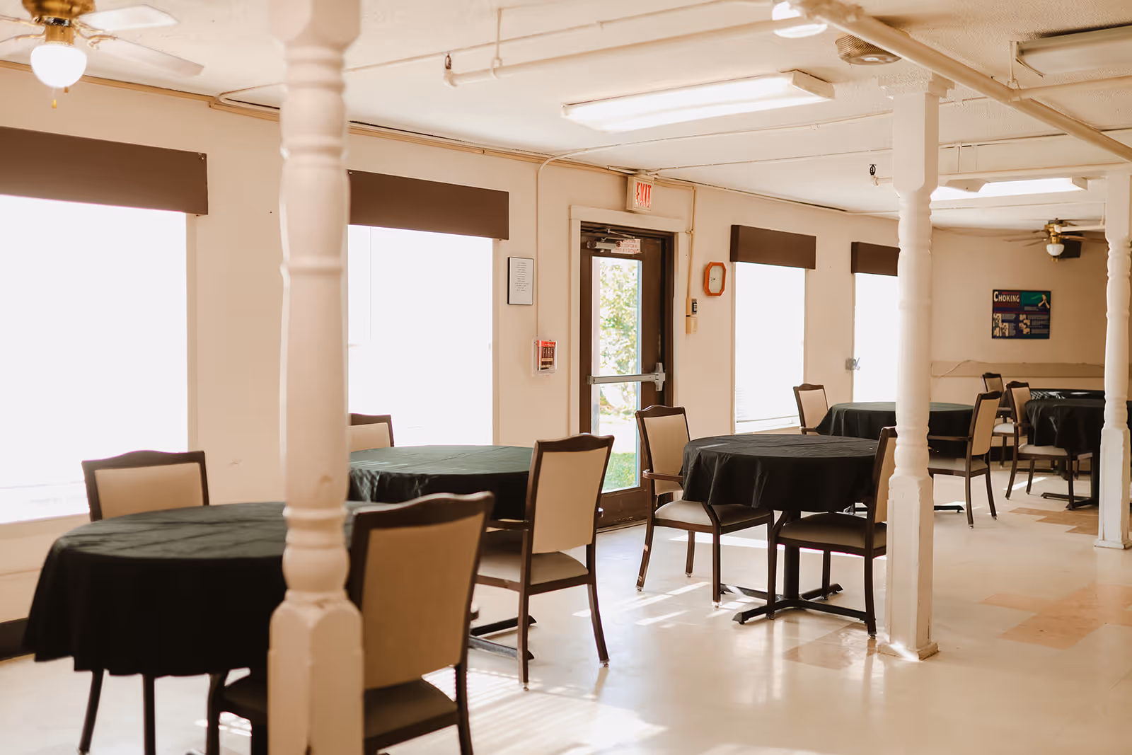 A dining room with several round tables covered with black tablecloths, each surrounded by beige cushioned chairs. The room has large windows with brown valances, white walls, white pillars, and a door leading outside. The floor is light-colored with some patterned tiles, and ceiling fans are visible.