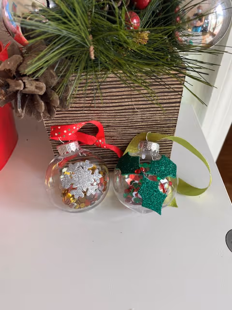 Two clear Christmas ornaments filled with glitter and confetti sit on a white surface next to pine greenery and a silver bauble.
