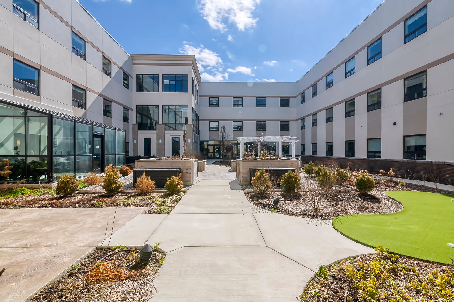 Outdoor courtyard area of The Avalon of Bloomfield Township senior living facility featuring a paved walkway, landscaped garden beds with shrubs, a white pergola, and a three-story building surrounding the courtyard under a partly cloudy blue sky.