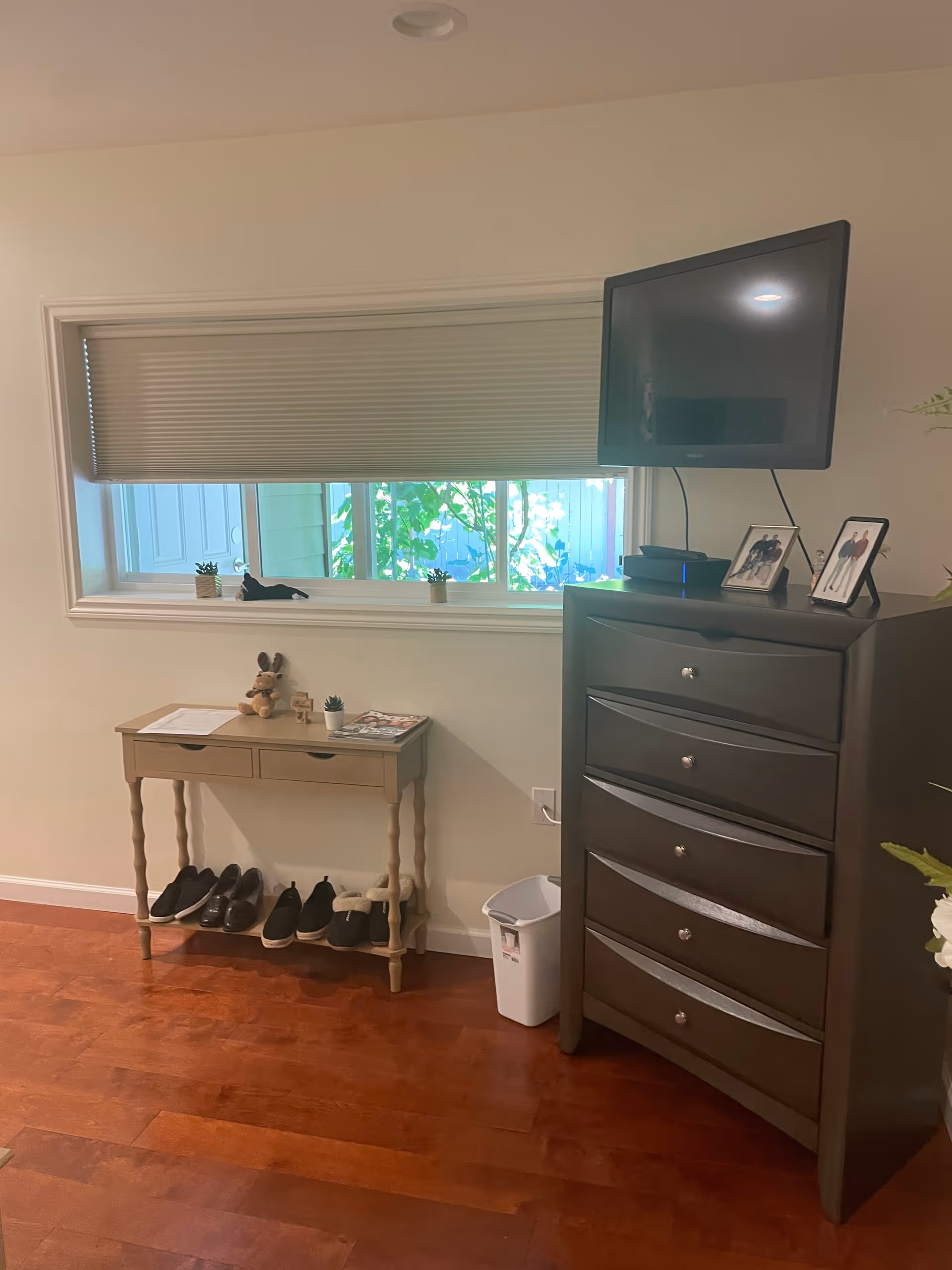 A room corner with a small wooden table holding a stuffed animal, a small plant, and some papers. Below the table are several pairs of shoes. Next to the table is a dark wooden chest of drawers with a flat-screen TV mounted above it and framed photos on top. A window with a partially closed blind shows greenery outside. The floor is wooden and there is a small white trash bin beside the chest of drawers.
