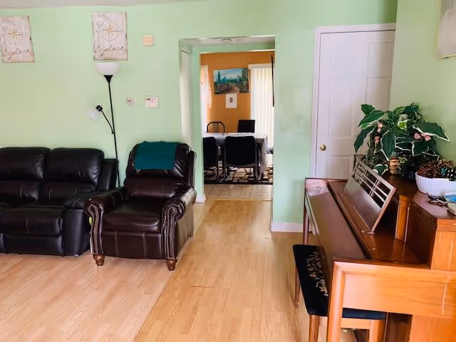 A living room area with light green walls, featuring a black leather sofa and armchair, a wooden piano with a bench, and a plant on top of the piano. In the background, a dining area with a table and chairs is visible through an open doorway.