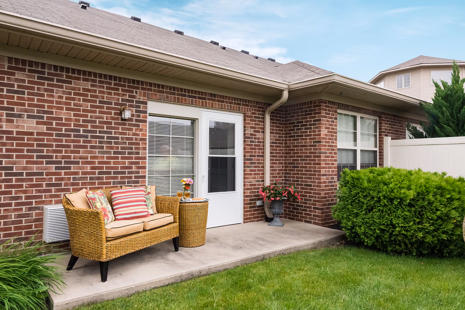 Brick patio outside a senior living residence featuring a wicker loveseat with colorful pillows, a small table, potted flowers, and a white door and windows.