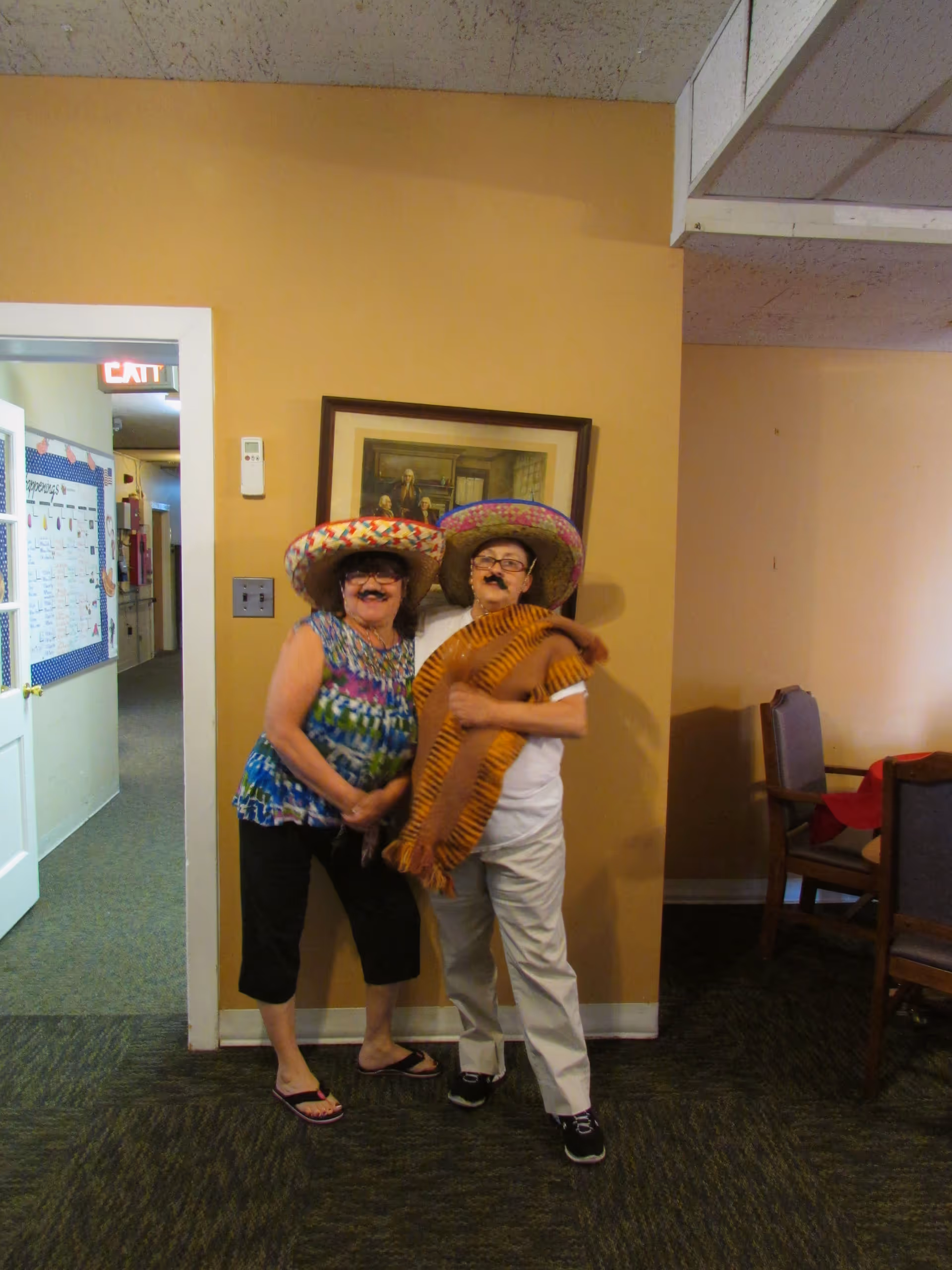 Two elderly women standing indoors against a yellow wall, wearing colorful sombreros and fake mustaches. One woman is dressed in a multicolored sleeveless top and black pants, while the other wears a white shirt, light-colored pants, and a striped shawl. There is a framed painting on the wall behind them and a doorway leading to a hallway on the left side of the image.