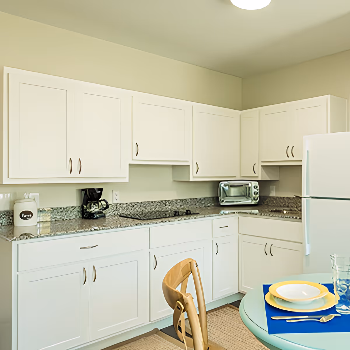 Bright kitchen with white cabinets, granite countertops, a refrigerator, toaster oven, and a small table set with a chair.