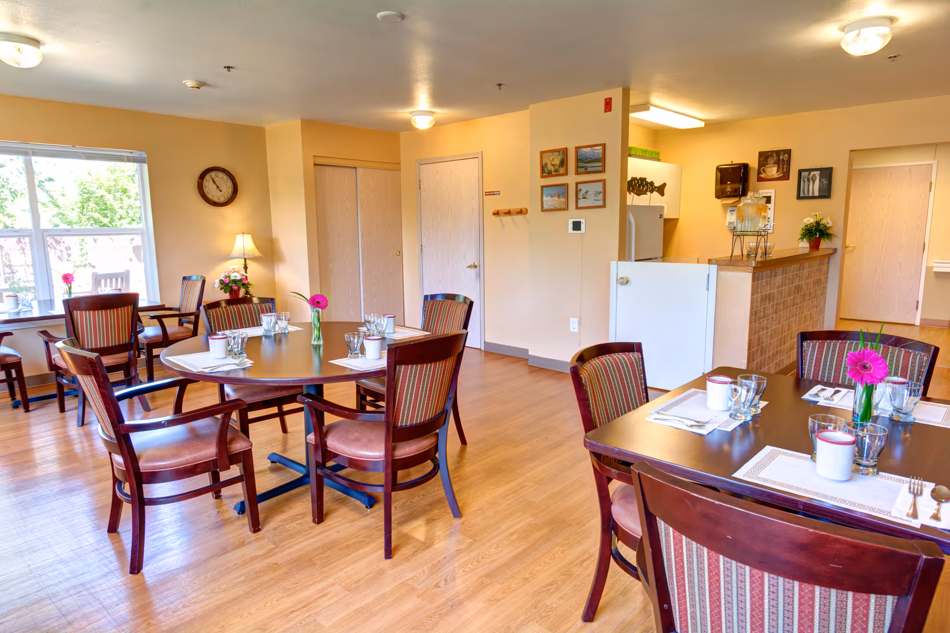 A bright dining room in a senior living facility with wooden tables and chairs arranged neatly. Each table is set with placemats, glasses, cups, and a small vase with a pink flower. The room has light-colored walls, a wooden floor, and a large window letting in natural light. There is a small kitchen area with a refrigerator and counter in the background, along with some framed pictures on the wall.