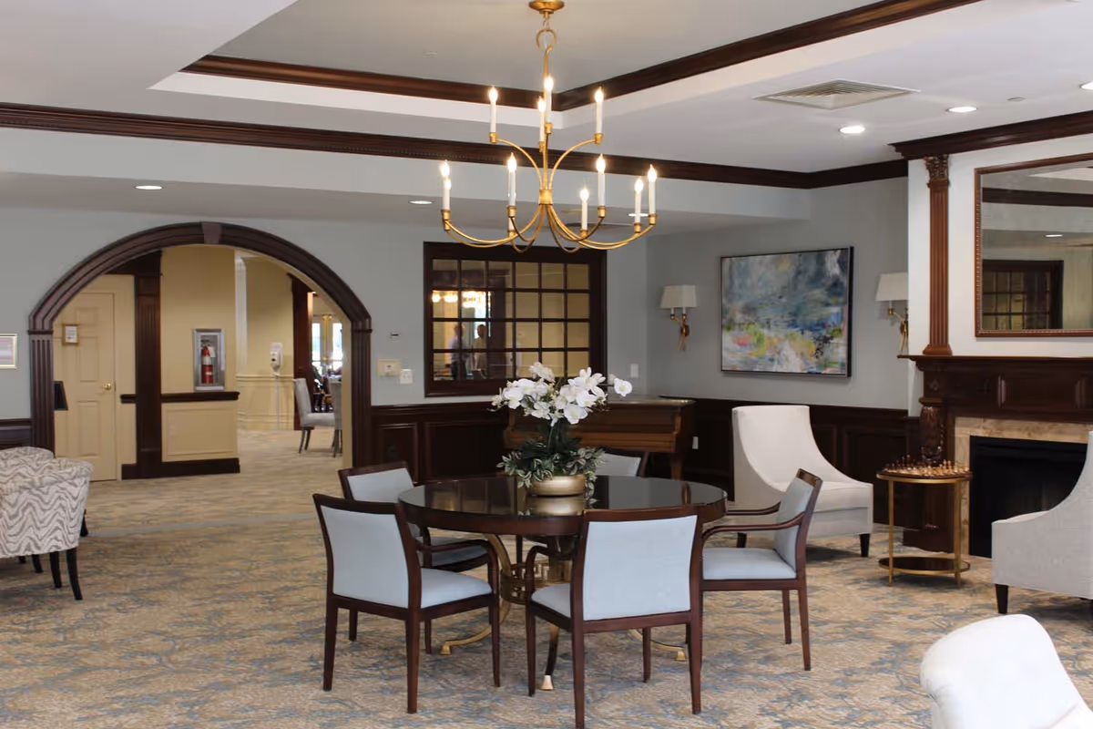 A spacious and elegant common area in a retirement community featuring a round wooden table with four upholstered chairs, a chandelier hanging from the ceiling, a fireplace with a large mirror above it, a side table with a chess set, and a floral arrangement on the table. The room has carpeted floors, light-colored walls with dark wood trim, and an arched doorway leading to another room.
