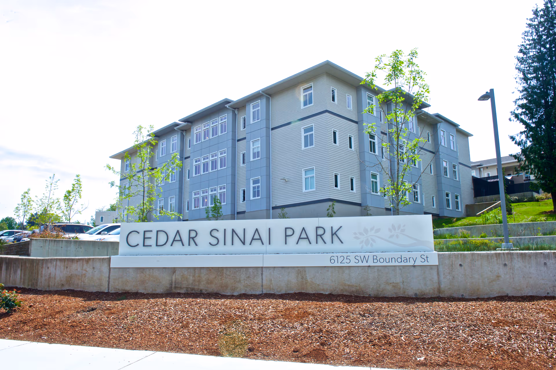 Exterior view of a multi-story residential building behind a large sign that reads 'CEDAR SINAI PARK' with the address '6125 SW Boundary St'. The building is surrounded by young trees and landscaping under a partly cloudy sky.