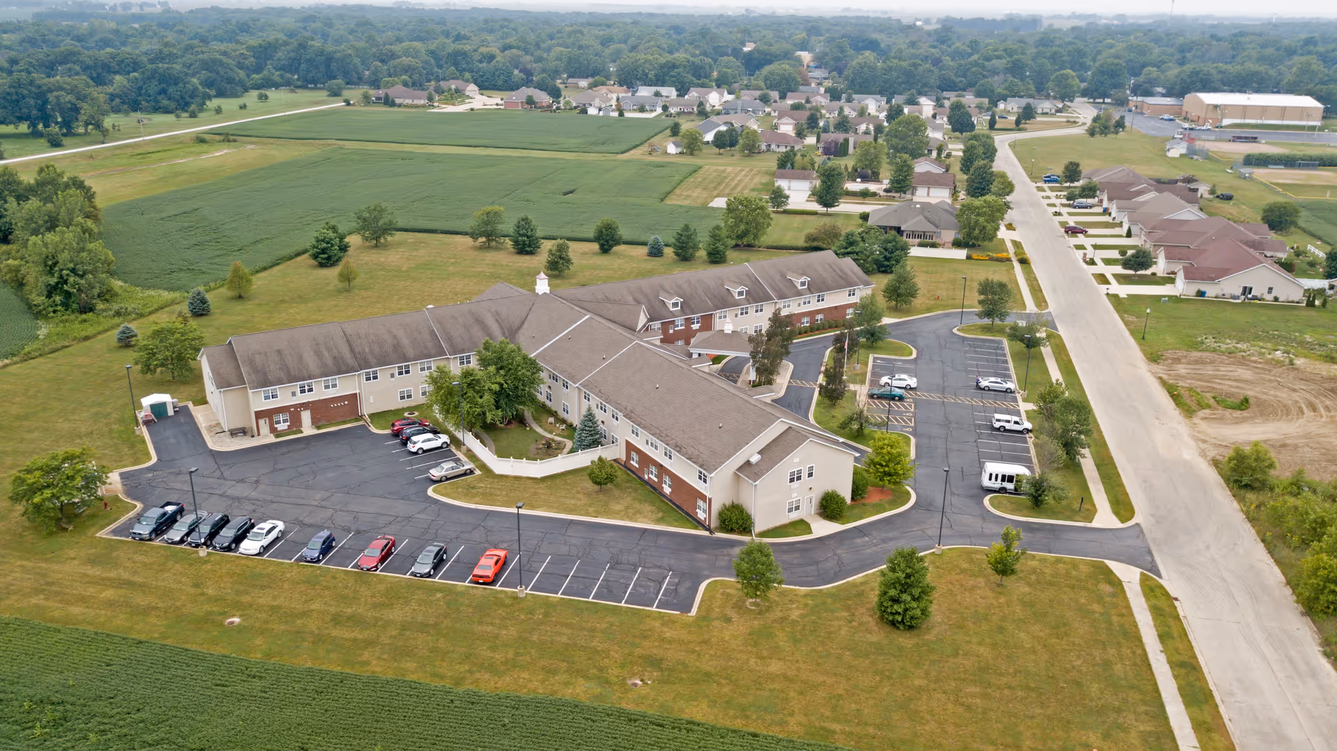 Aerial view of Heritage Woods of Watseka, a senior living facility surrounded by green fields and a residential neighborhood. The building is a large, two-story structure with a parking lot containing several cars. The facility is situated in a rural area with open spaces and trees around it.