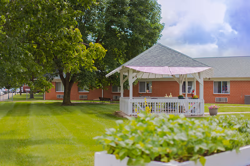 A white gazebo on a manicured lawn with trees in front of a single-story red-brick building.
