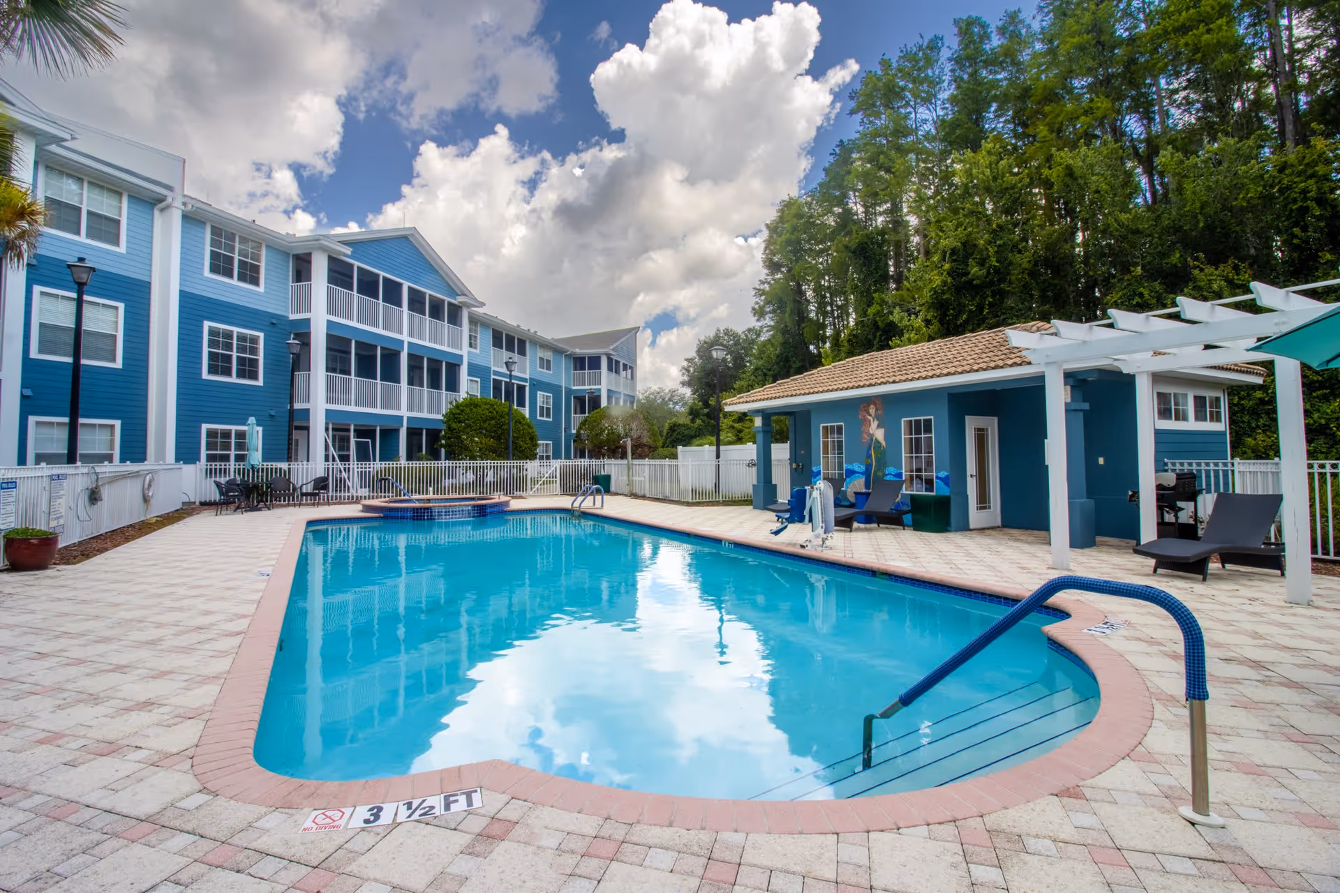 Outdoor swimming pool area at Addington Place of East Lake with a clear blue pool, poolside chairs, a small building with a tiled roof, and a blue multi-story residential building in the background under a partly cloudy sky.