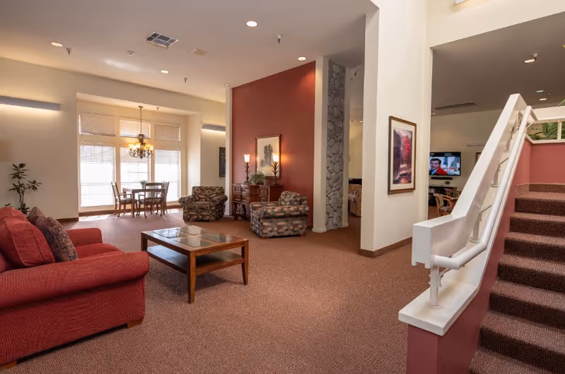 Spacious senior living common area with sofas, armchairs, a coffee table, dining nook, and a carpeted staircase.