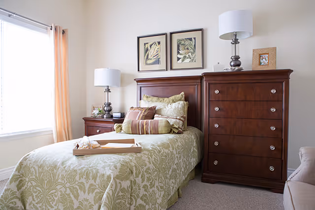 A neatly made single bed with green and beige patterned bedding and multiple pillows in a bright bedroom. The bed has a wooden headboard and a tray with a cloth on it. There are two matching wooden nightstands on either side of the bed, each with a lamp. A wooden dresser with drawers and a framed photo on top is next to the bed. Two framed botanical prints hang on the wall above the headboard. A window with light curtains allows natural light into the room.