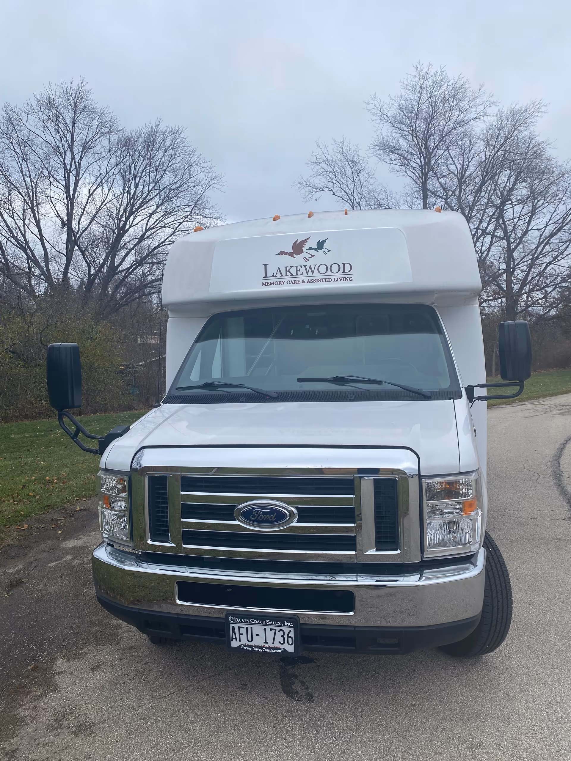 Front view of a white Lakewood Memory Care & Assisted Living shuttle van parked on a road showing the Ford grille and license plate.
