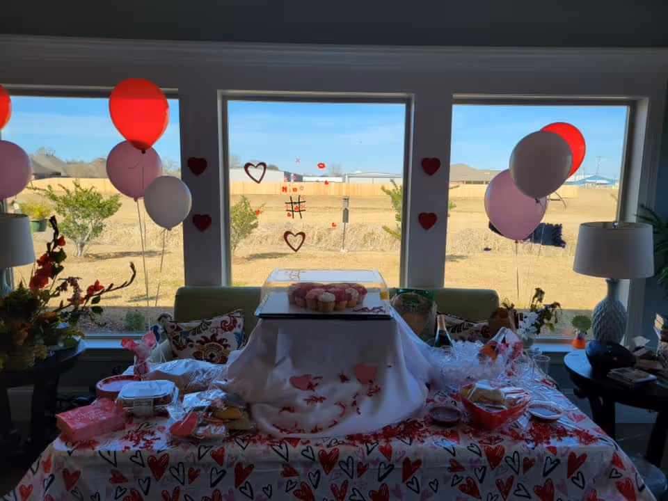 A festive table set up in front of three large windows with a view of a backyard. The table is covered with a heart-patterned tablecloth and decorated with red, pink, and white balloons. There are cupcakes under a clear cover, various snacks, and drinks on the table. Heart-shaped decorations are stuck on the windows.