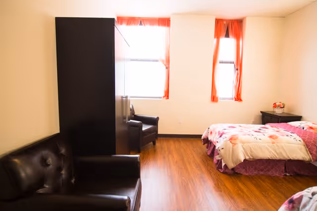 A bright senior living bedroom with two windows covered by orange curtains, a bed with floral bedding, a black leather armchair, a black leather sofa, a black wardrobe, and a small nightstand with a flower vase.