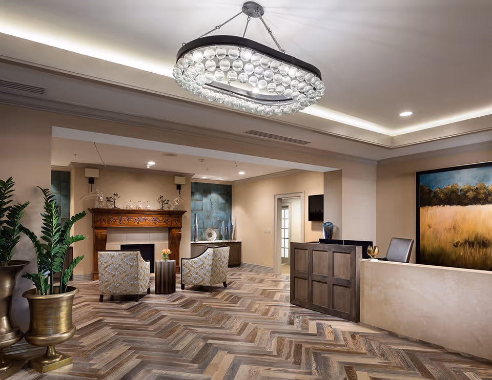 A stylish reception lobby with a chandelier, seating area, decorative fireplace, potted plants, and a wooden reception desk on herringbone floors.