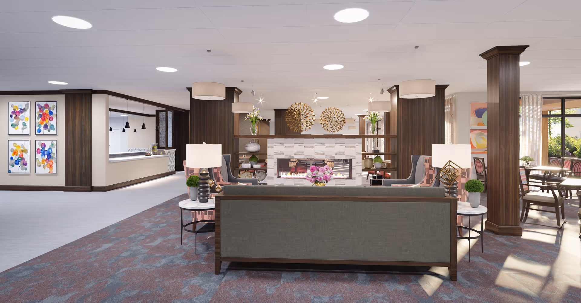 A spacious and well-lit senior living facility common area featuring a modern fireplace with decorative golden wall art above it. The room includes comfortable seating with a gray sofa and patterned armchairs, side tables with lamps and small plants, and large windows allowing natural light to fill the space. The area has wood-paneled columns, colorful abstract artwork on the walls, and a dining area visible in the background.