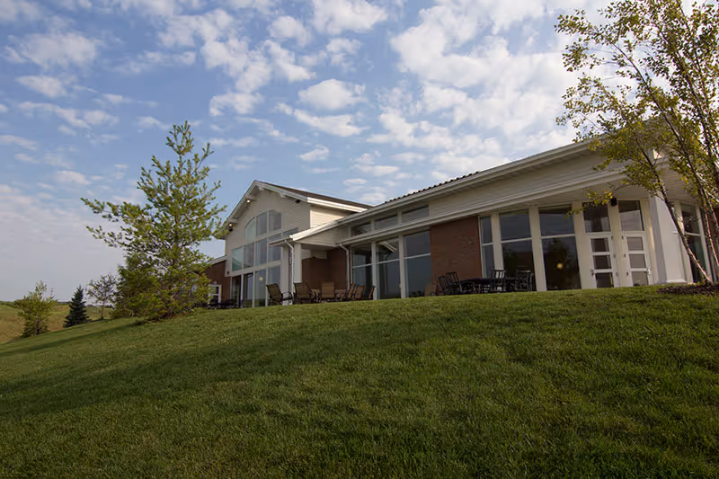 Exterior view of a single-story building with large windows and a patio area with chairs, situated on a grassy hill under a partly cloudy sky.