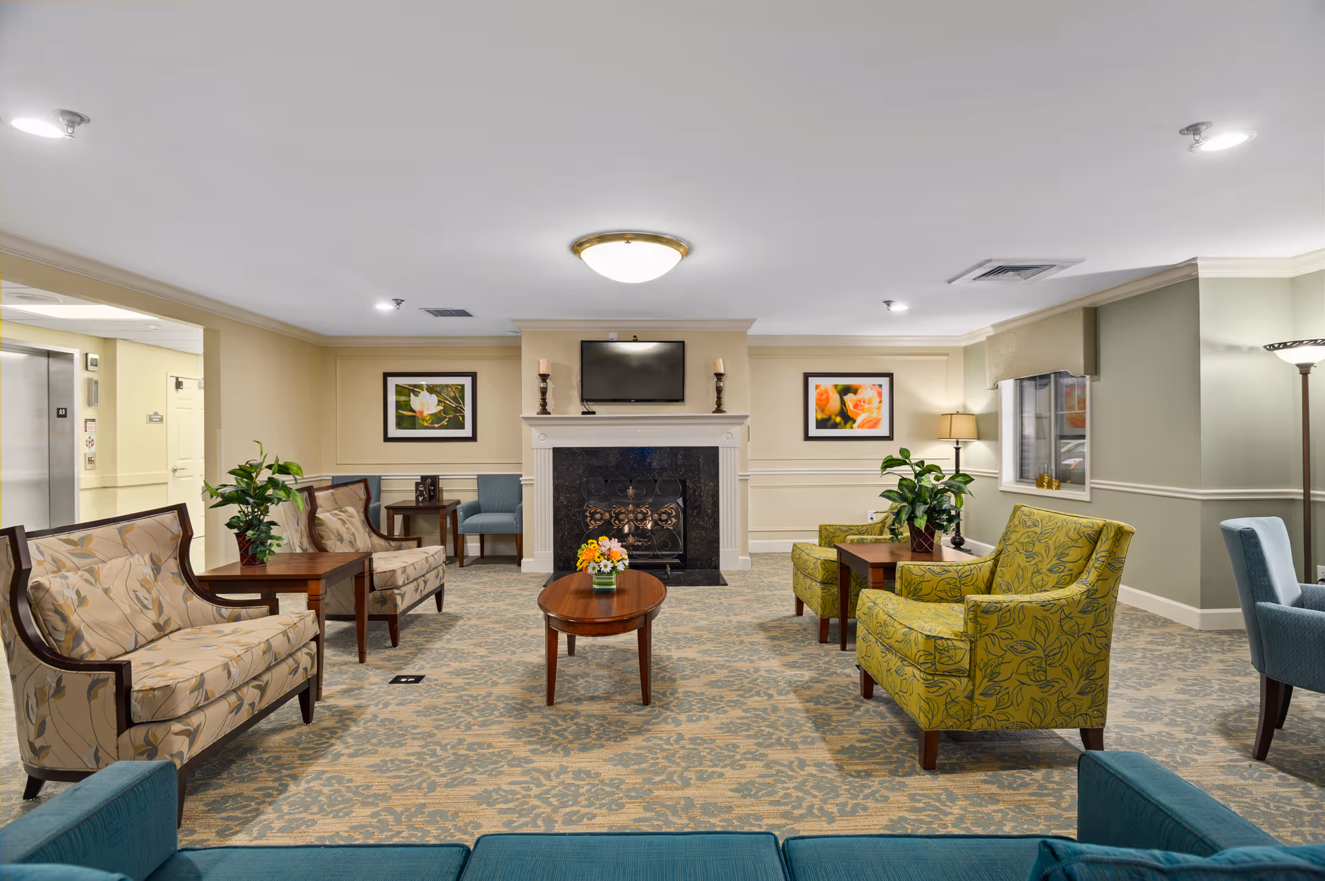 A spacious senior living facility common area with patterned carpet, multiple upholstered chairs and sofas in beige, green, and blue tones, wooden coffee tables with flower arrangements, a fireplace with a TV mounted above it, framed floral artwork on the walls, and soft lighting from ceiling fixtures and lamps.