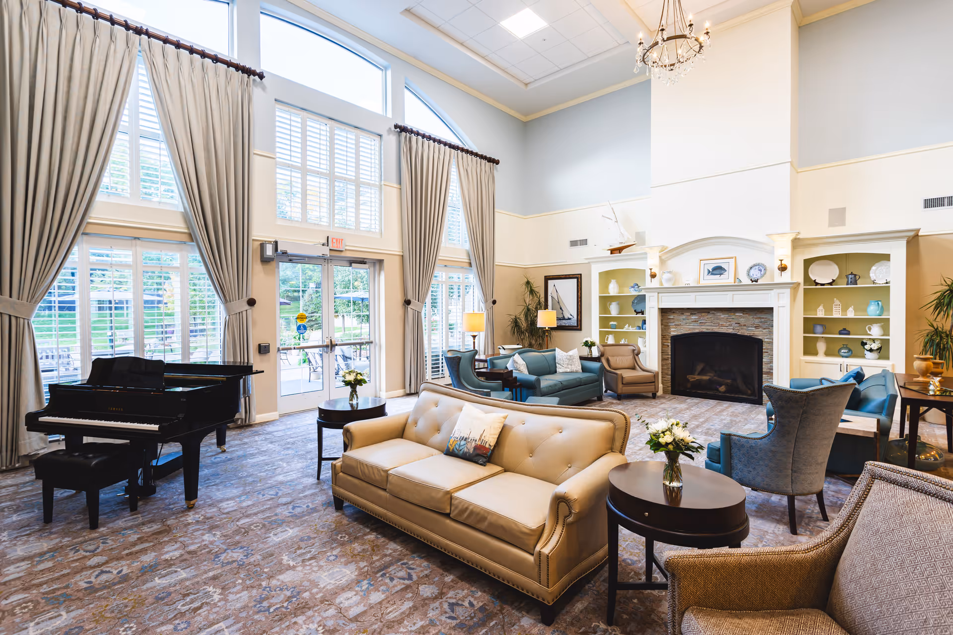 A spacious and elegant living room with high ceilings and large windows covered by long curtains. The room features a beige sofa, multiple armchairs, a grand piano, a fireplace with decorative shelves on either side, and several small tables with flower arrangements. The room is well-lit with natural light and a chandelier hanging from the ceiling.