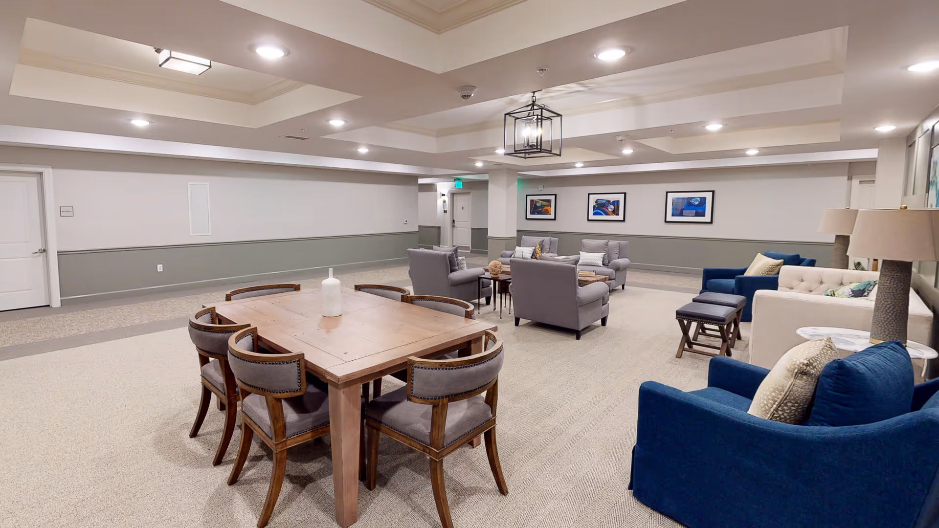 A spacious, well-lit common area with a wooden table surrounded by six chairs in the foreground. The background features multiple cushioned armchairs and sofas arranged around small tables. The walls are decorated with framed artwork, and the ceiling has recessed lighting and a central hanging light fixture.