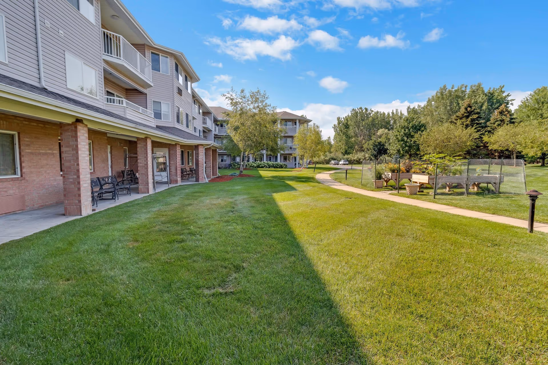 Outdoor view of Independence Village of Saginaw showing a multi-story residential building with balconies and a covered patio area with benches. The foreground features a well-maintained grassy lawn and a paved walking path that curves through the landscaped grounds with trees and garden beds under a partly cloudy blue sky.