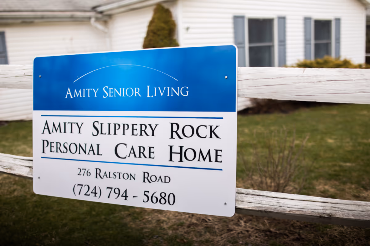 A sign attached to a wooden fence in front of a white building with windows. The sign reads 'Amity Senior Living, Amity Slippery Rock Personal Care Home, 276 Ralston Road, (724) 794-5680'.
