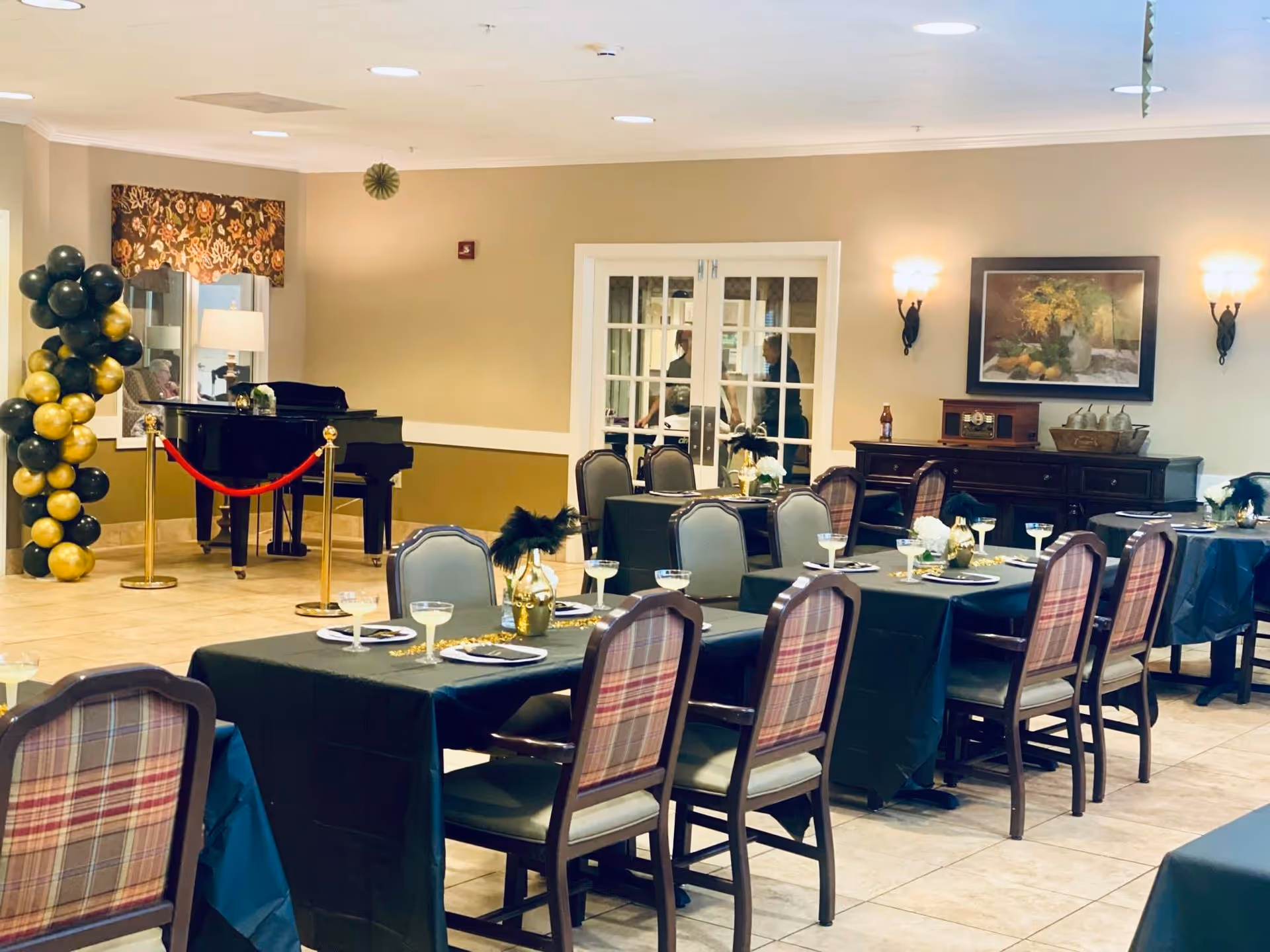 A dining room set up with tables covered in black tablecloths, each table decorated with gold vases holding black feathers and set with plates and glasses. The room features plaid upholstered chairs, a grand piano roped off with a red velvet rope, a balloon arrangement in black and gold, and a sideboard with decorative items and a framed painting on the wall.